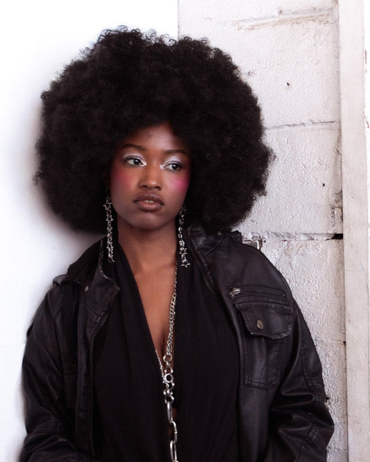 Woman with large afro wearing a black jacket, star earrings, and an aria chain against a white brick wall