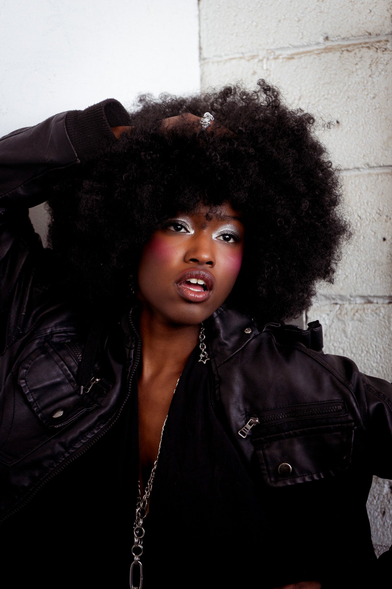 Woman with large afro wearing a black jacket, star earrings, and an aria chain against a white brick wall