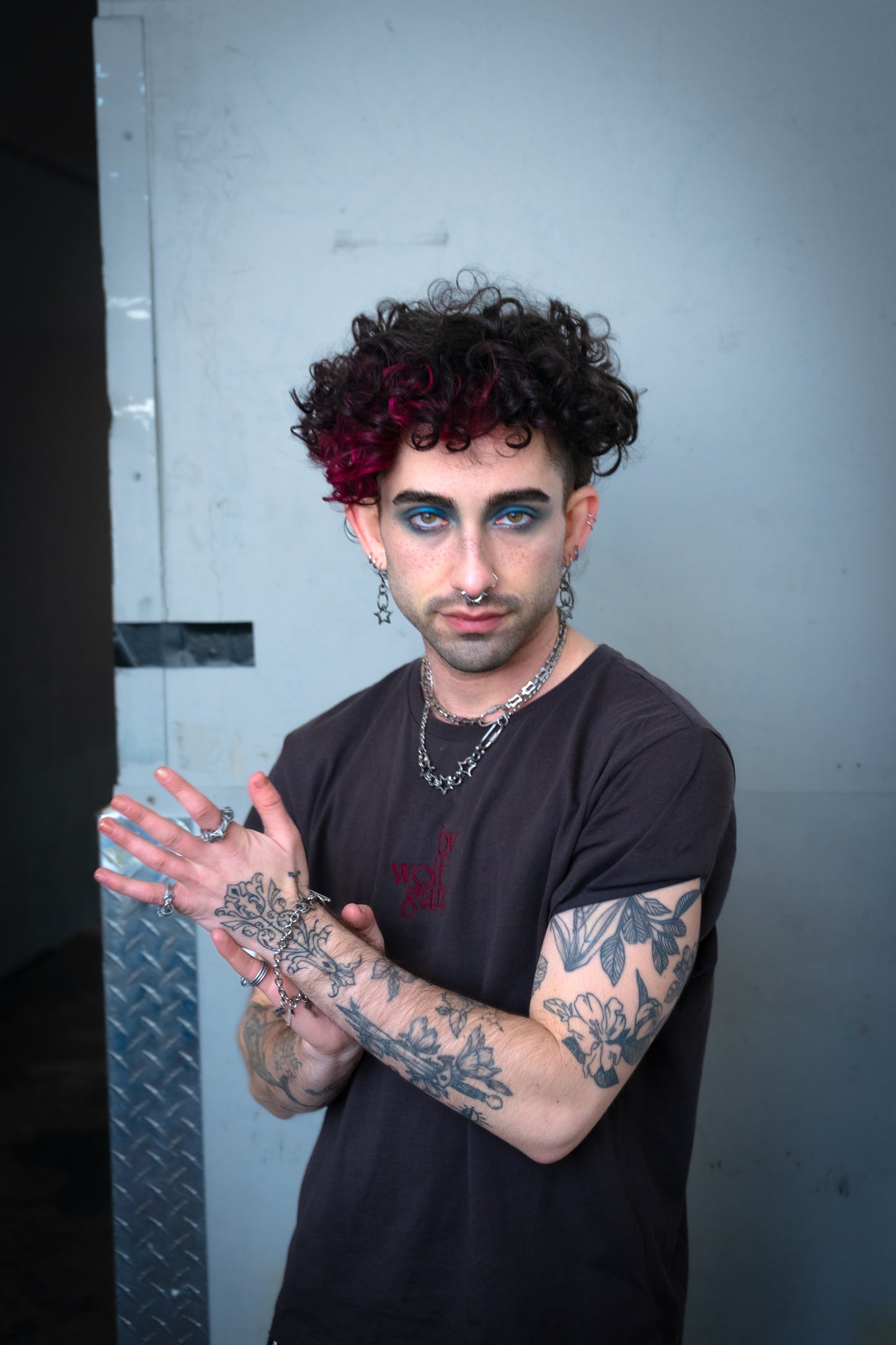 Person with tattoos and dark hair posing against a gray wall wearing bywolfgang earrings, rings, and necklaces.