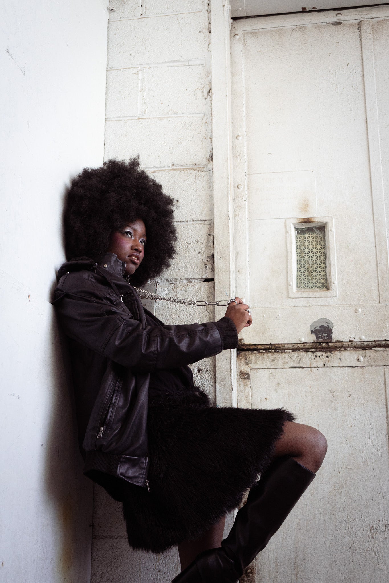 Woman with large afro wearing a black jacket, star earrings, and an aria chain with boots, chained to a door in a dimly lit room. 
