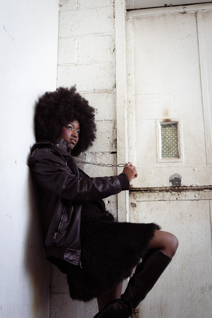 Woman with large afro wearing a black jacket, star earrings, and an aria chain with boots, chained to a door in a dimly lit room. 