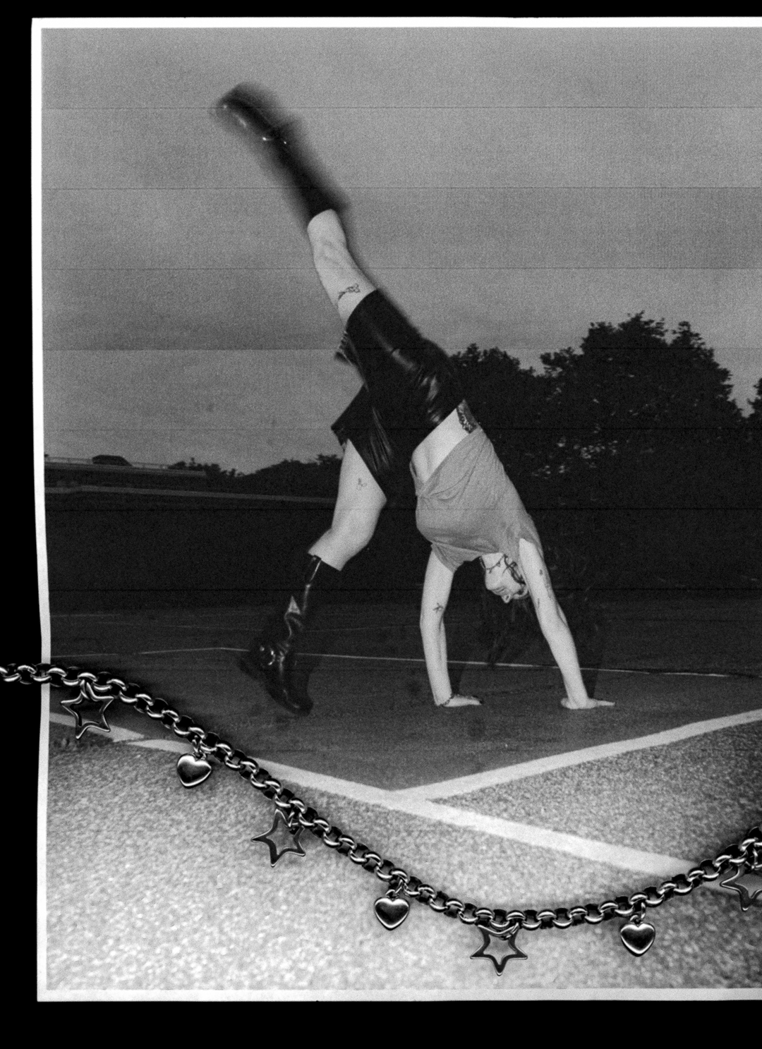 Person doing a handstand on a tennis court with a charm necklace with hearts and hollow star charms in lsck and white photocopy edit scan. 