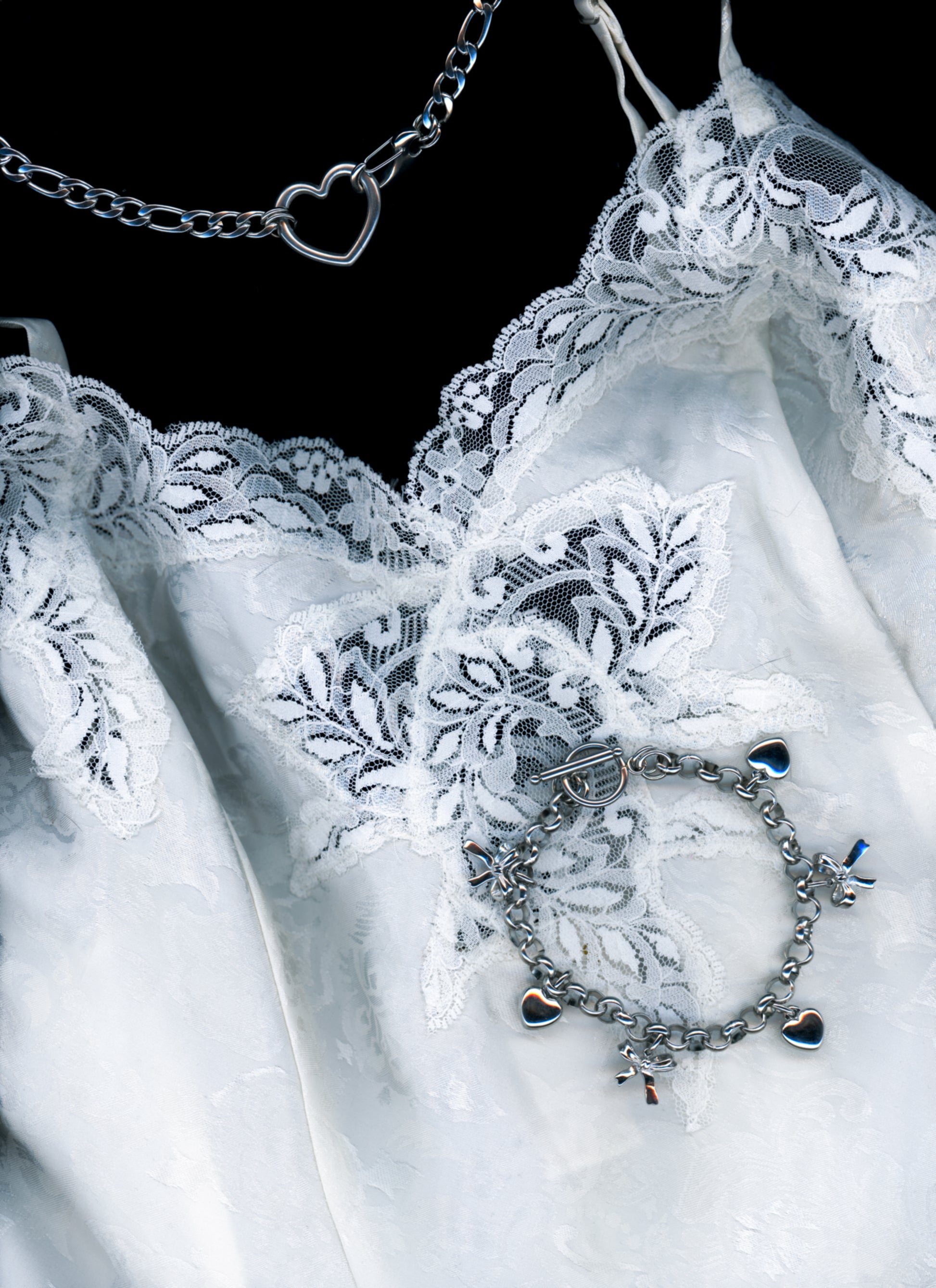 a silver Figaro chain necklace with a heart shaped closure, and a charm bracelet with 3-d bow and heart charms re displayed draped on a white satin and lace slip on a black background