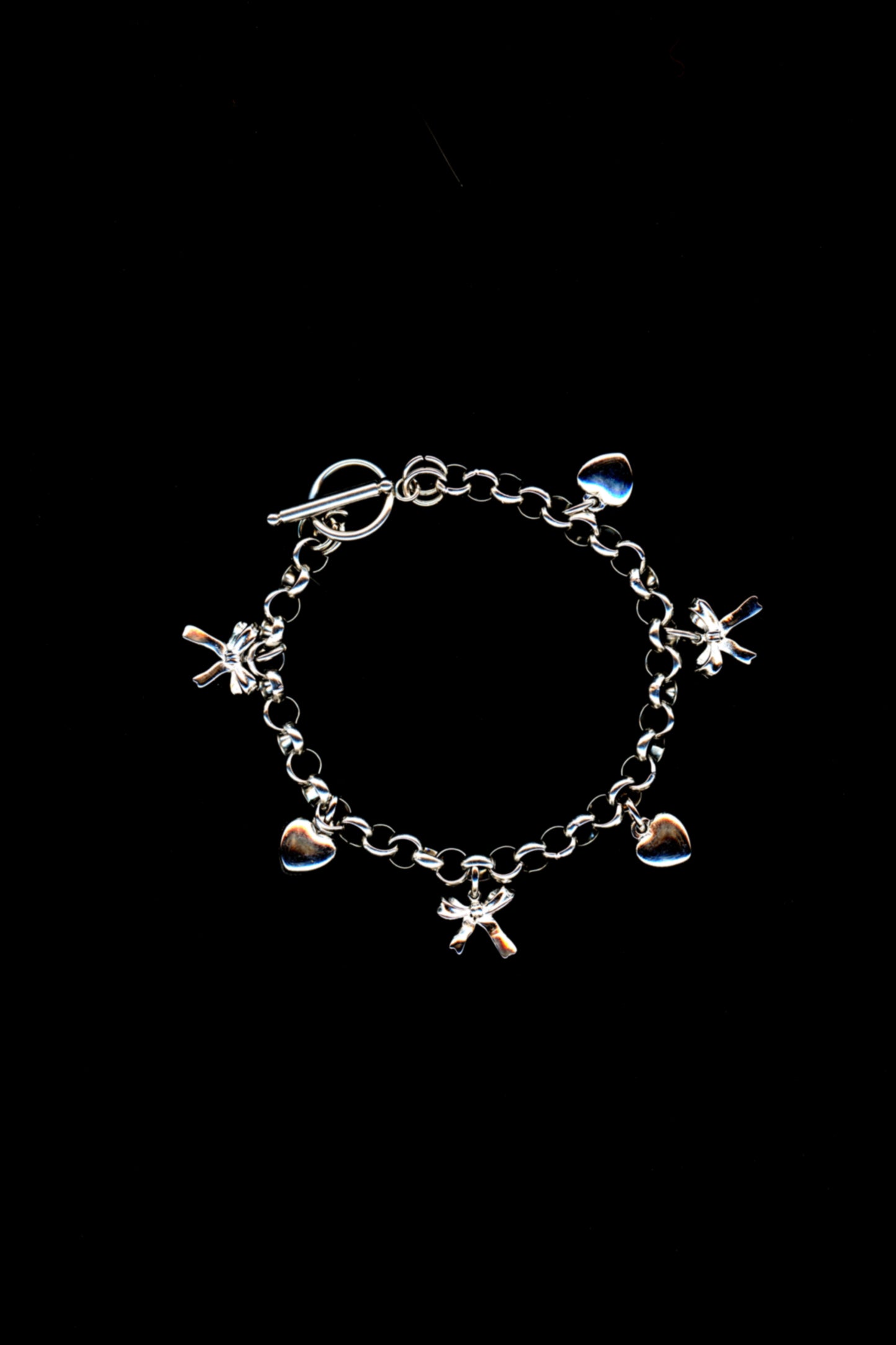 A silver charm bracelet with a three-dimensional bow and heart charms, and toggle closuredisplayed against a black background. 