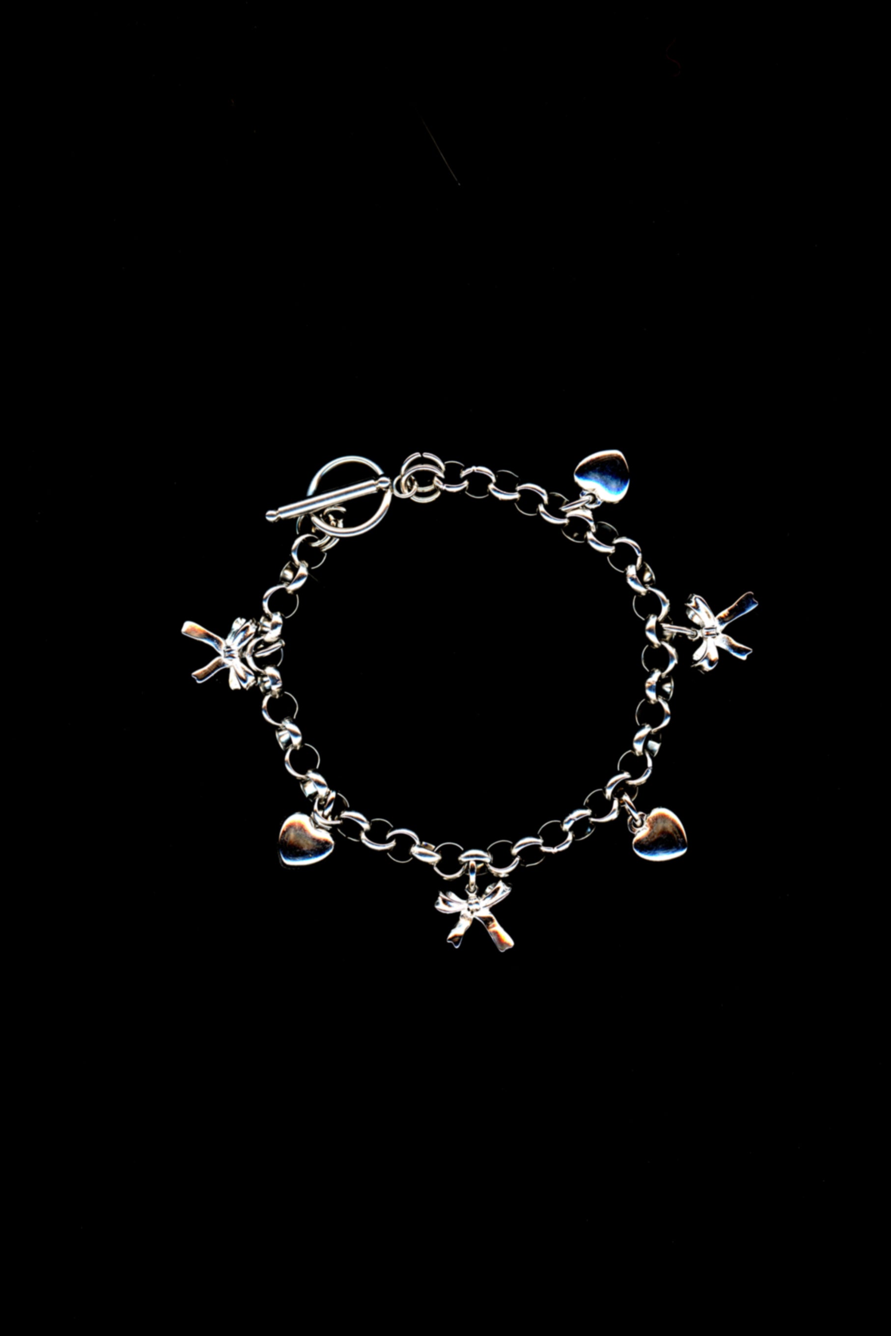 A silver charm bracelet with a three-dimensional bow and heart charms, and toggle closuredisplayed against a black background. 